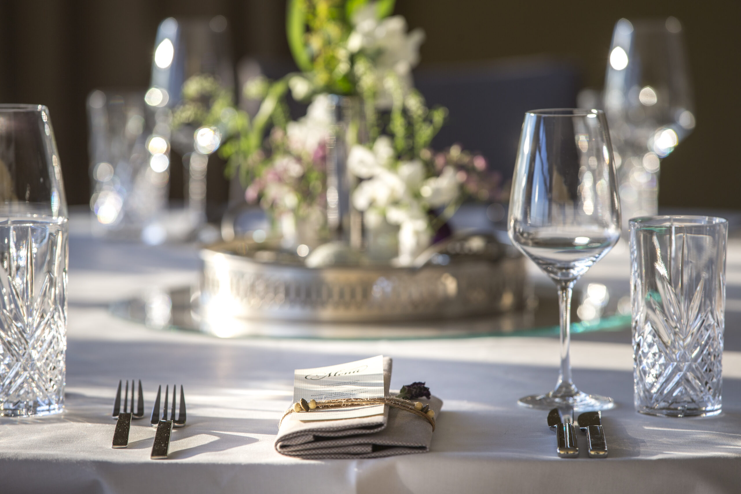 Detailansicht gedeckter Tisch zu einem feierlichen Anlass mit Serviette, Weinglass und Besteck. Im Hintergrund sieht man verschwommen Blumen